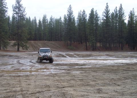 1990 Ford Bronco - Jeep Wrangler navigates a muddy off-road course under an overcast sky.
