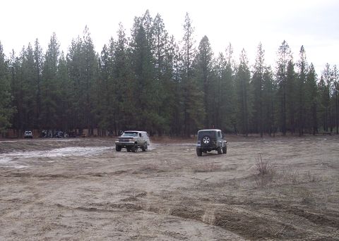 1990 Ford Bronco - Two off-road vehicles navigate a dirt track surrounded by a dense forest.