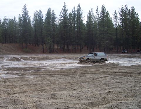 1990 Ford Bronco - A gray Ford Bronco navigates a muddy terrain with trees lining the horizon.