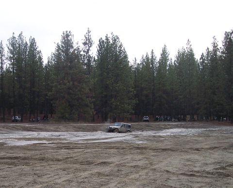 1990 Ford Bronco - A white SUV navigates a muddy terrain with a line of trees in the distance and other vehicles parked.