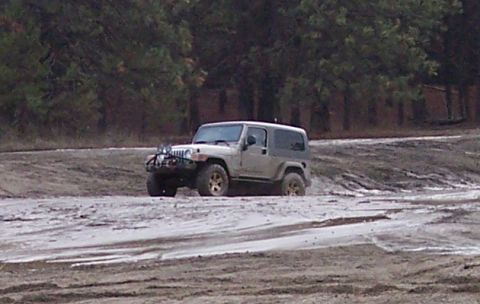 1990 Ford Bronco - A Jeep Wrangler TJ navigates a muddy terrain with trees visible behind it.