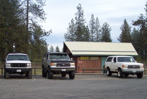 1990 Ford Bronco - Three Ford Broncos are parked in a lot near a building and trees.