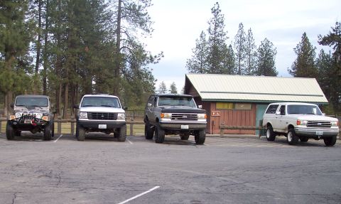 1990 Ford Bronco - Four-wheel drive vehicles are parked in a lot near a building with trees in the background.