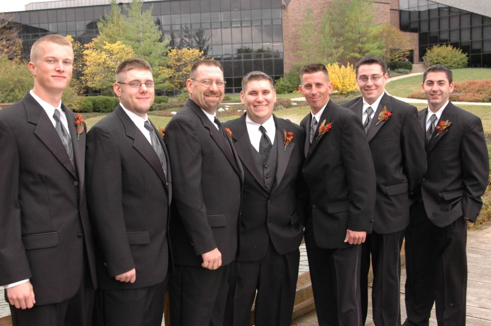 Seven men in black tuxedos stand together outdoors