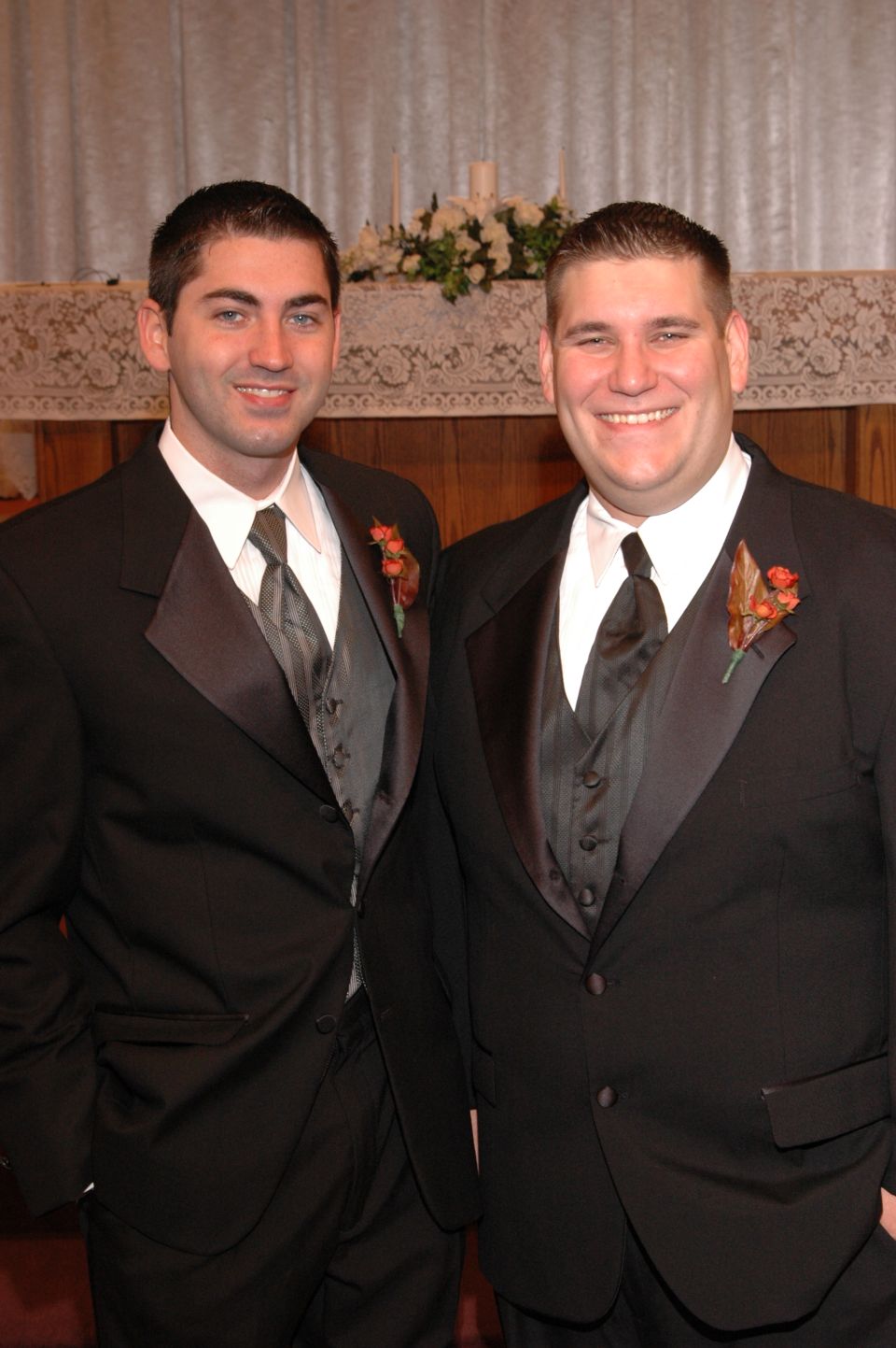 Two men in black tuxedos with grey vests and ties stand smiling