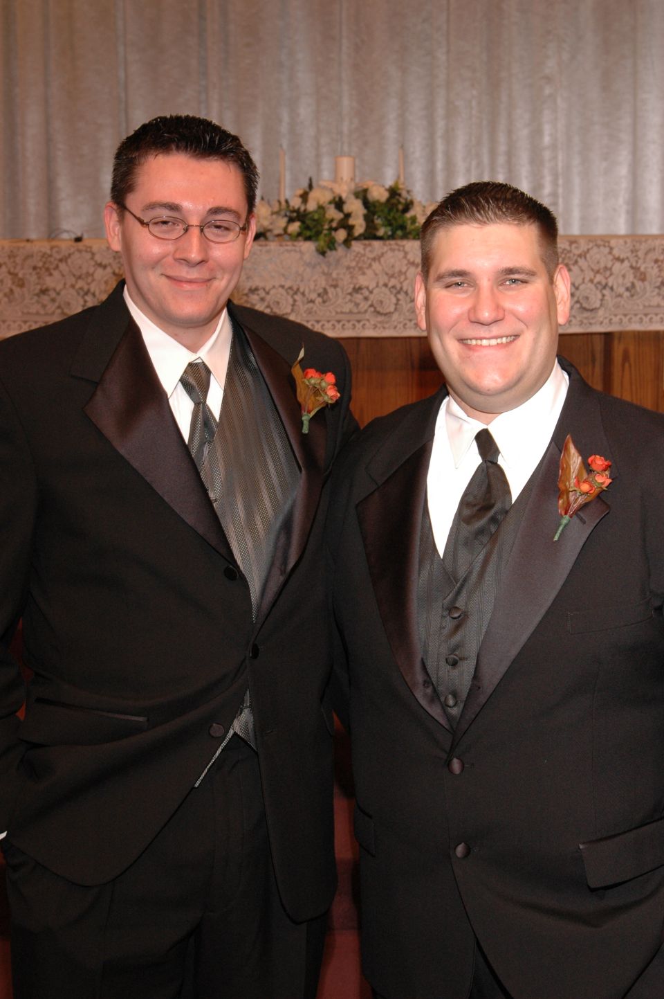 Two men in black tuxedos with boutonnieres stand in front of a lace-covered altar