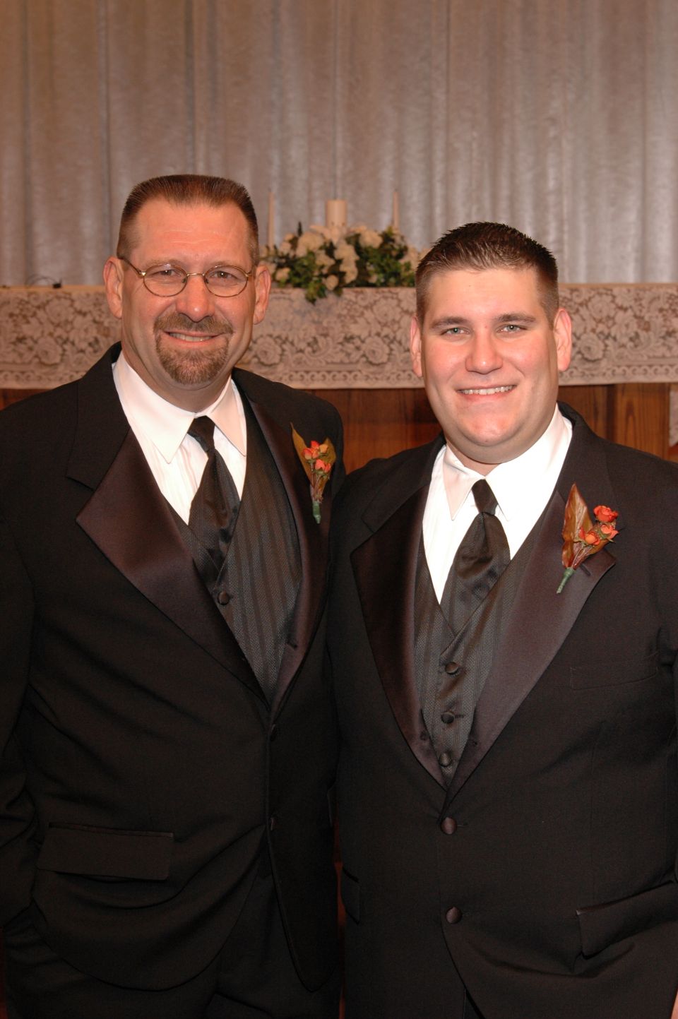 Two men in tuxedos stand together, smiling. The man on the left wears glasses and has a goatee