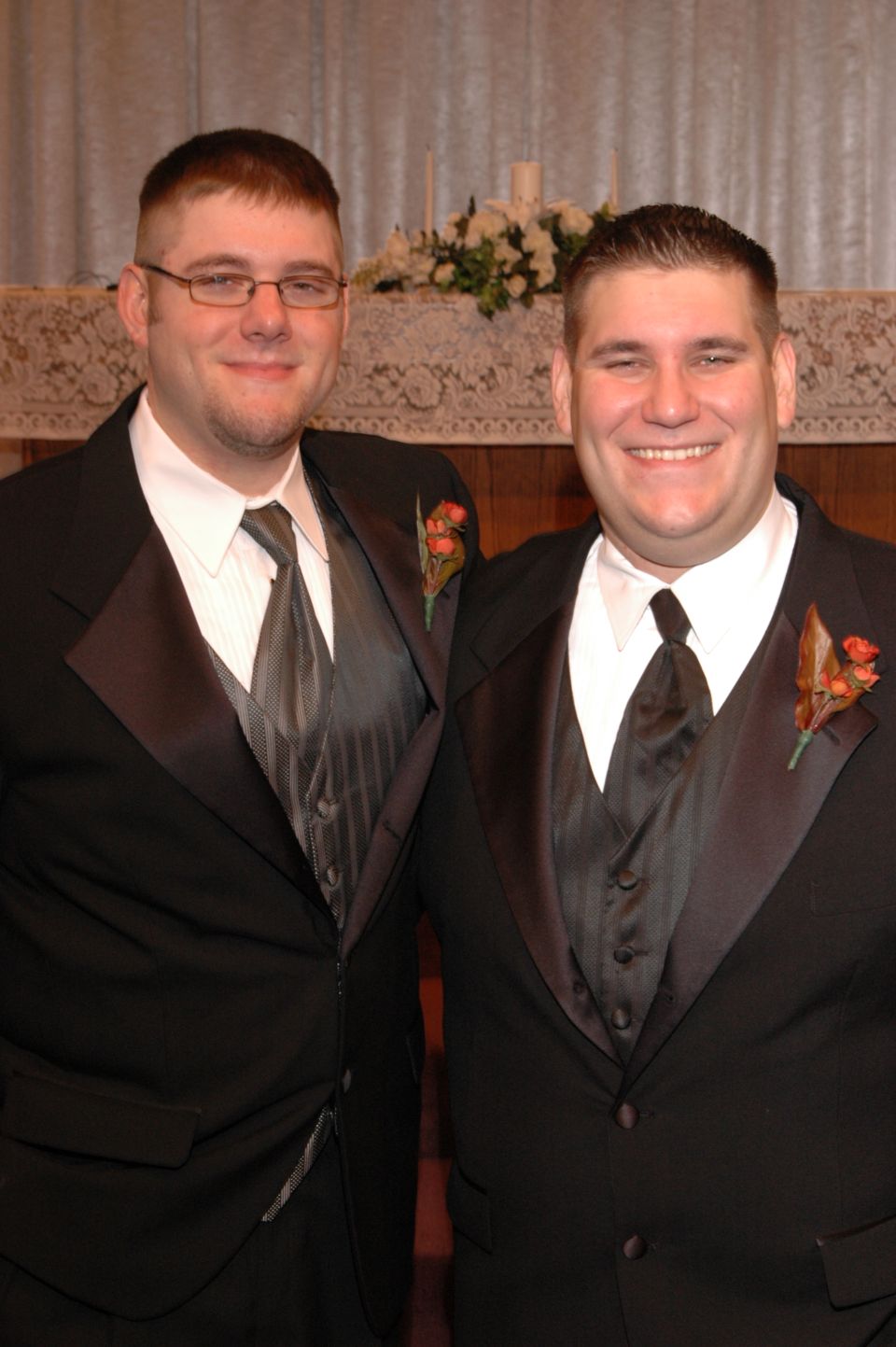 Two men in formal wear, one with glasses, stand together. A floral arrangement is behind them