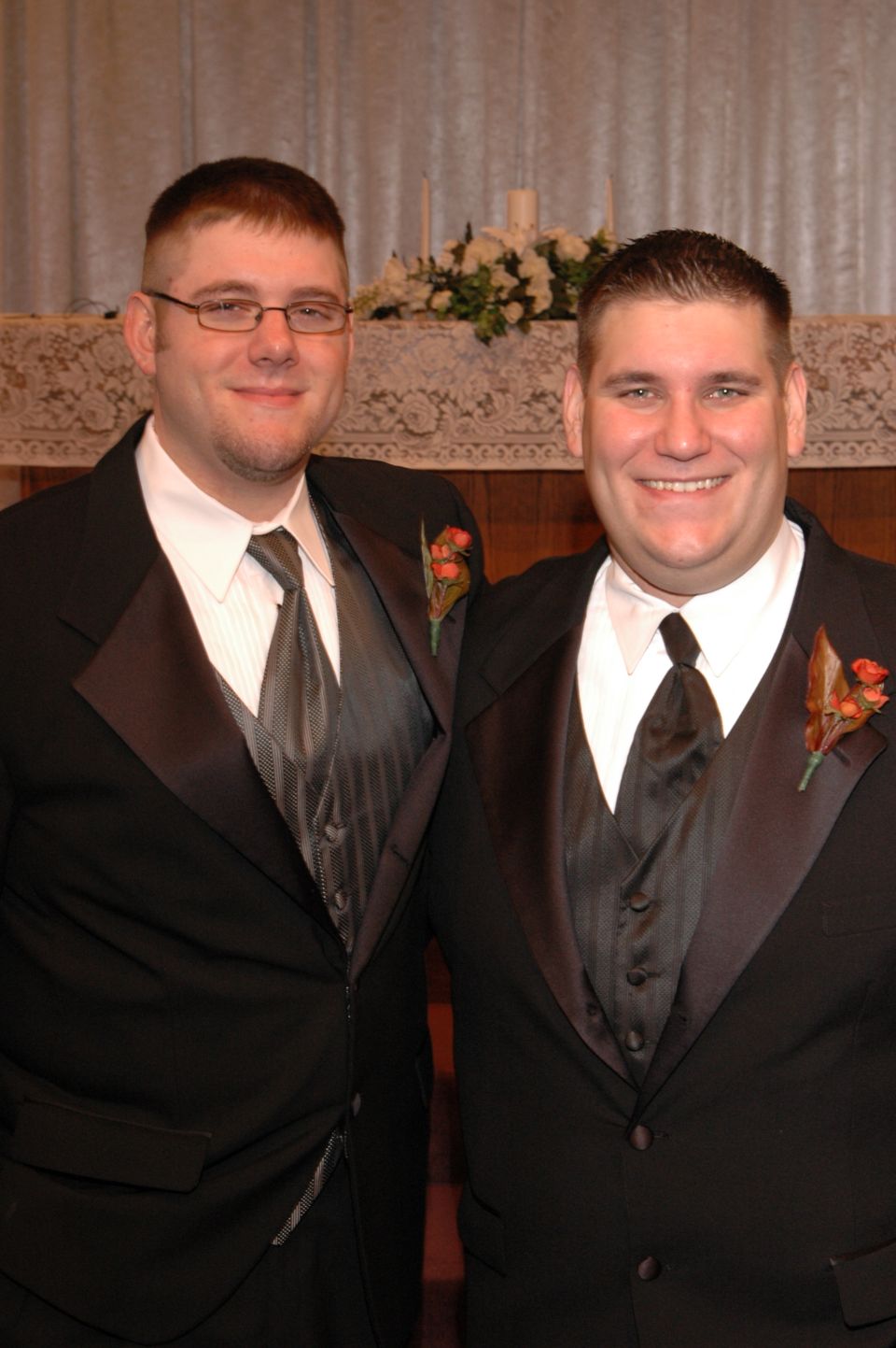 Two men in tuxedos stand together, smiling. They both have boutonnieres