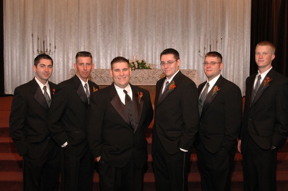 Six men in black tuxedos with grey ties stand in front of a white curtain