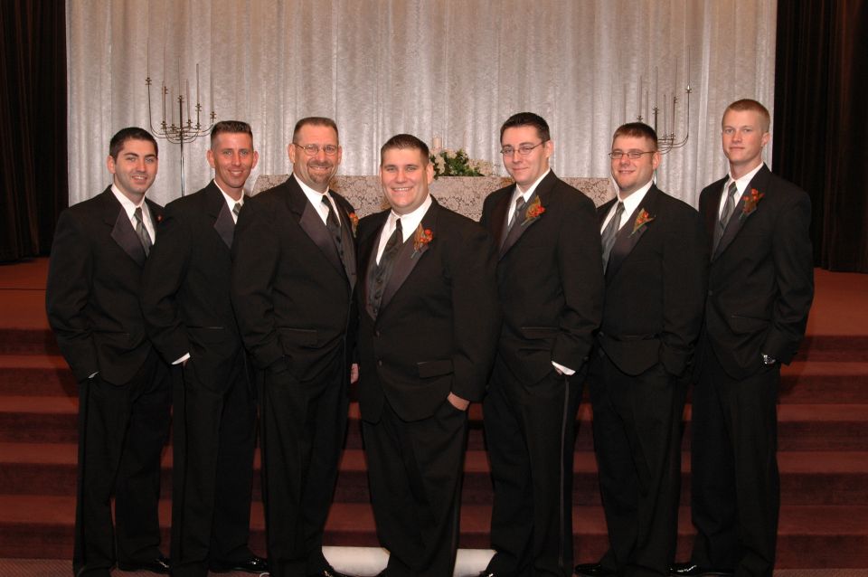 Seven men in black tuxedos with orange boutonnieres stand in a line on red carpeted steps