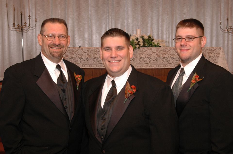 Three men in black tuxedos with ties and boutonnieres stand smiling in front of a white curtain and altar