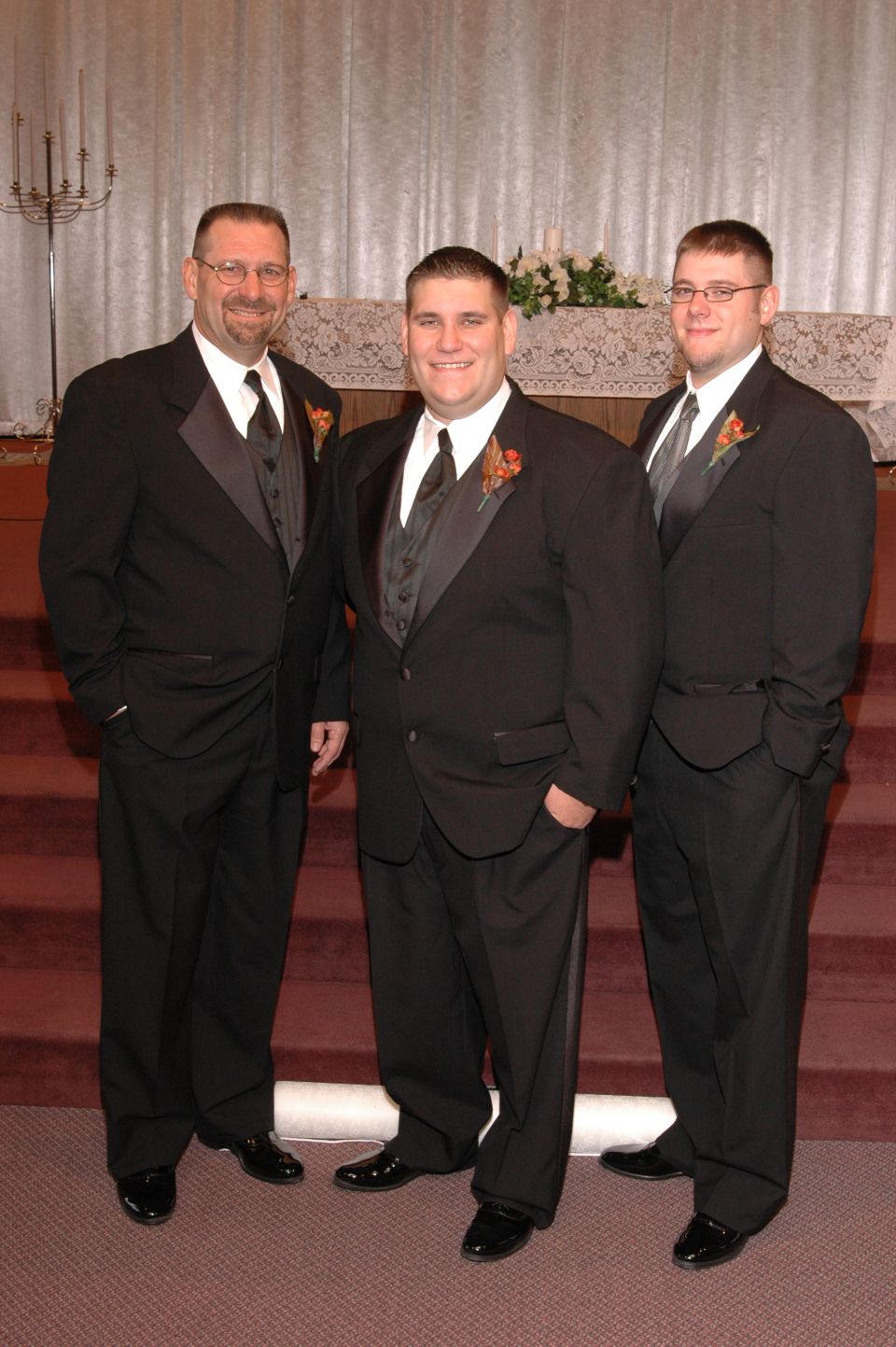 Three men in black tuxedos stand on red carpeted steps in front of a white curtain