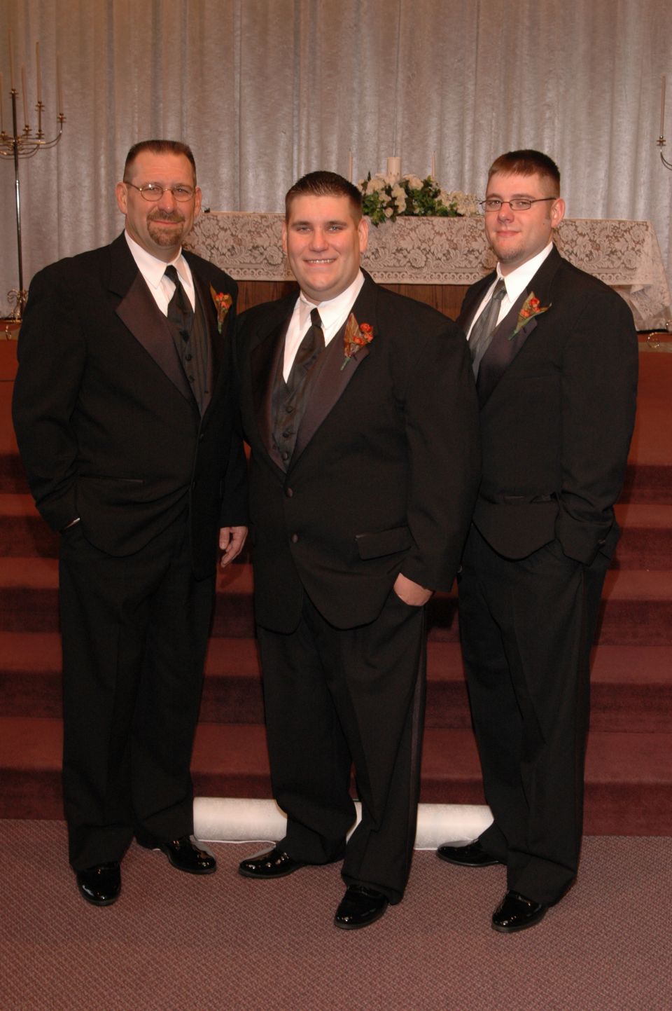 Three men in black tuxedos stand in front of a decorated altar