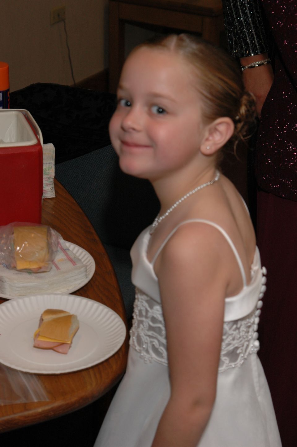 Girl in a white dress with a pearl necklace smiles at the camera. Sandwiches are on plates on a table to her left