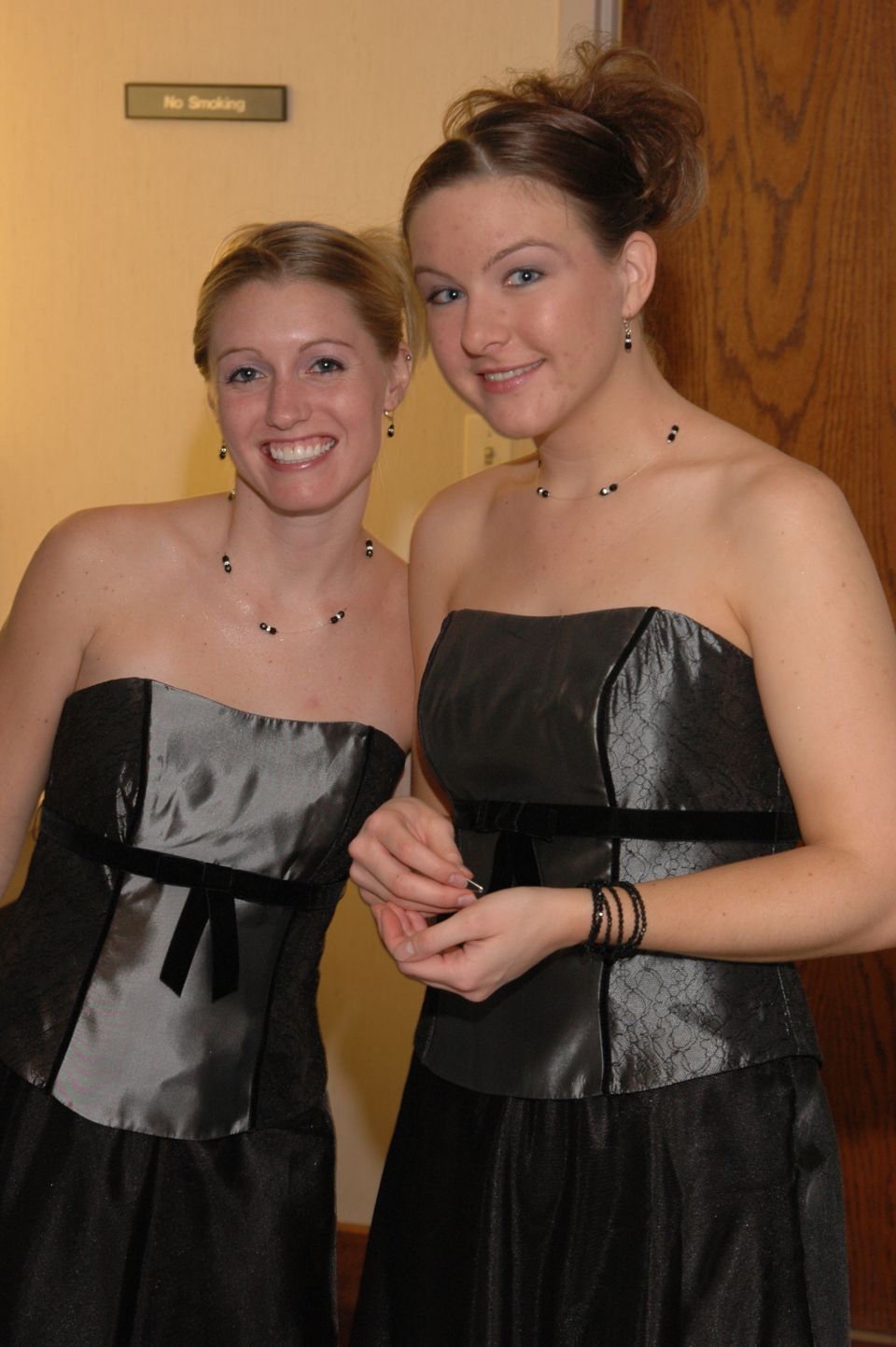 Two women in strapless black dresses stand indoors. A 'No Smoking' sign is visible on the wall