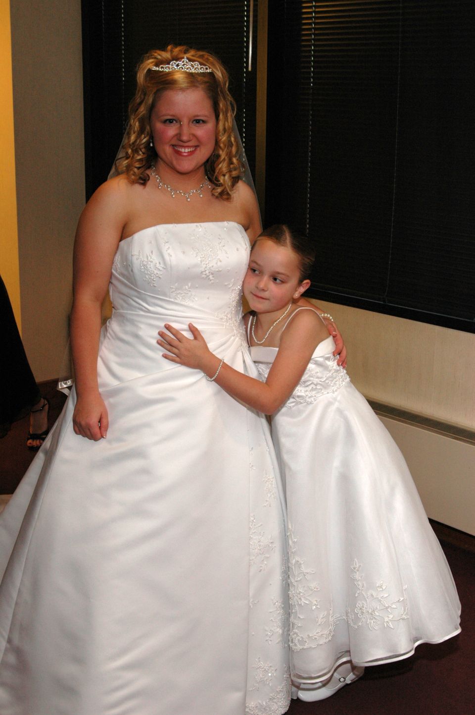 Bride in a white strapless gown and tiara hugs a young flower girl in a matching white dress
