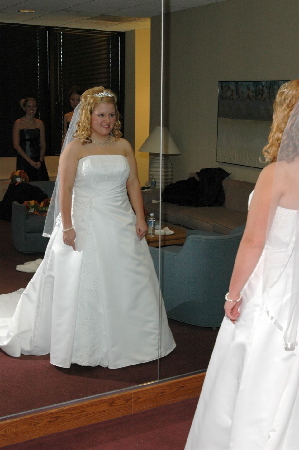 Bride in a white strapless wedding dress with a tiara and veil, looking at her reflection in a large mirror