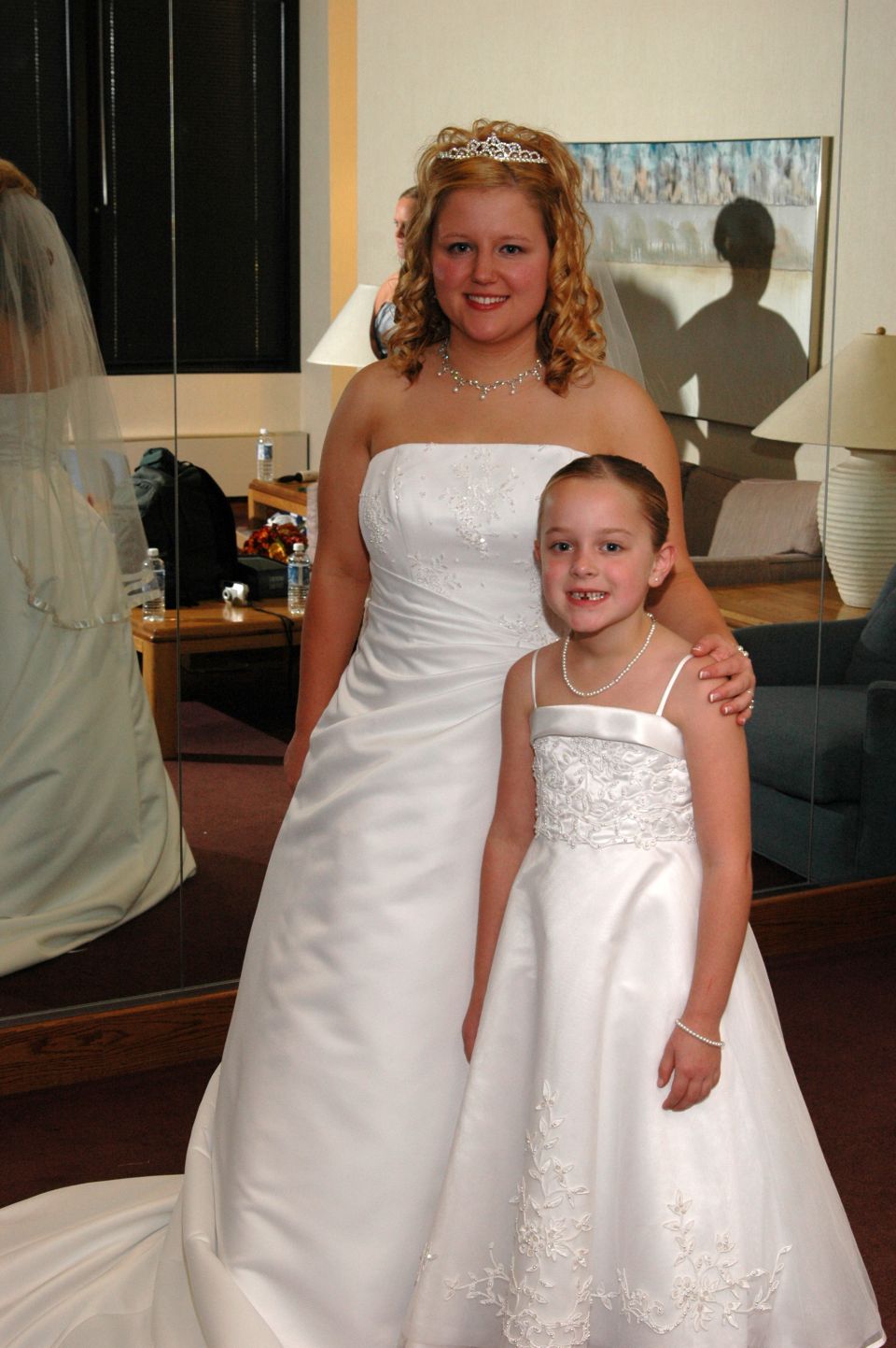 Bride and flower girl in white dresses stand in front of a mirror