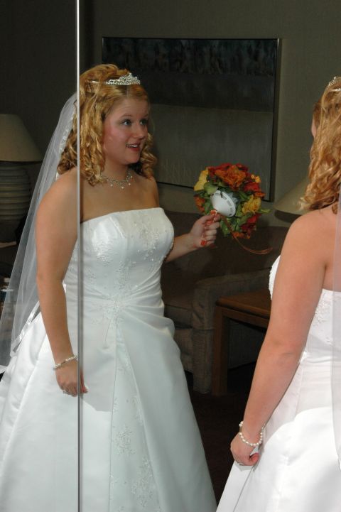 1985 Ford Crown Victoria LTD - A bride in her wedding attire smiles as she looks at her reflection in a large mirror, holding a colorful bouquet.