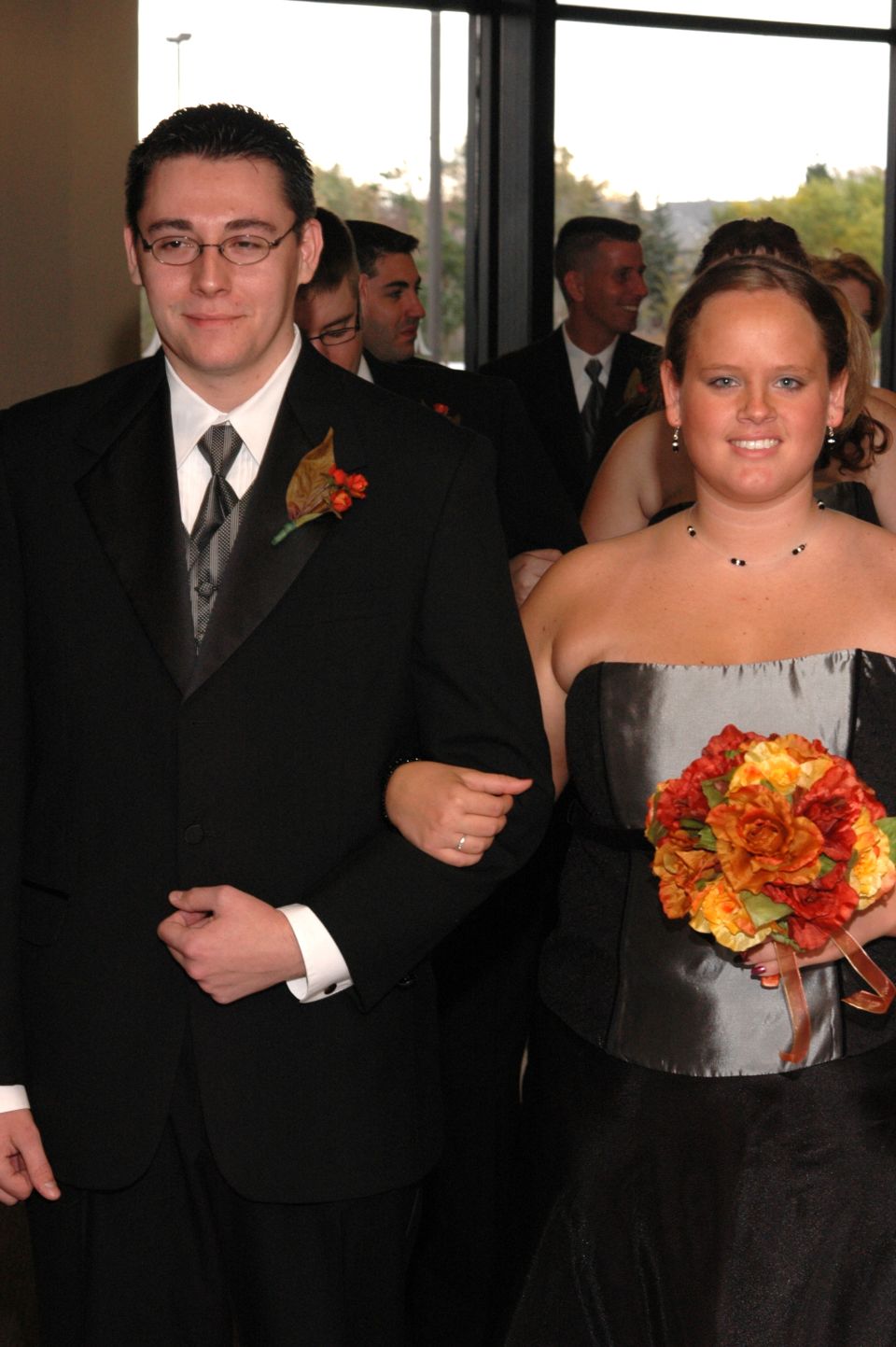 Wedding party members walking together, man in tuxedo arm-in-arm with woman in formal dress holding flowers