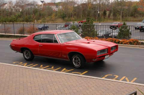 1985 Ford Crown Victoria LTD - A bright red 1969 Pontiac GTO is parked in a lot with other vehicles in the background.