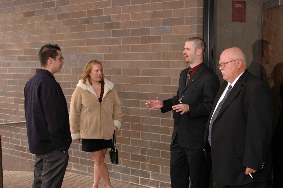 People in formal attire stand outside a brick building, with one man gesturing