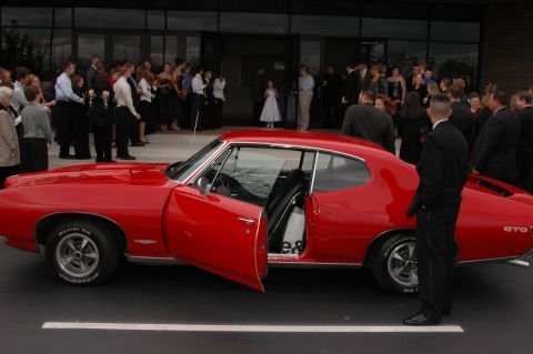 1985 Ford Crown Victoria LTD - A classic red Pontiac GTO is parked in front of a building where a gathering of people are present.