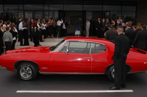 1985 Ford Crown Victoria LTD - A bright red 1967 muscle car is parked in front of a building where a formal event is taking place.