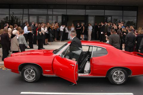 1985 Ford Crown Victoria LTD - A red 1967 Pontiac GTO is parked near a building entrance where wedding guests are gathered.
