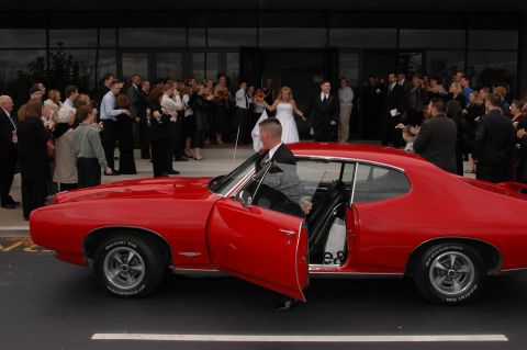 1985 Ford Crown Victoria LTD - A man in a suit exits a classic red coupe as a wedding party gathers outside a building.