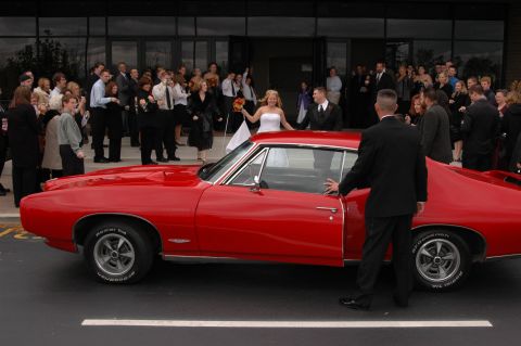 1985 Ford Crown Victoria LTD - A red 1969 Pontiac GTO is ready for a wedding, with guests gathered in the background.