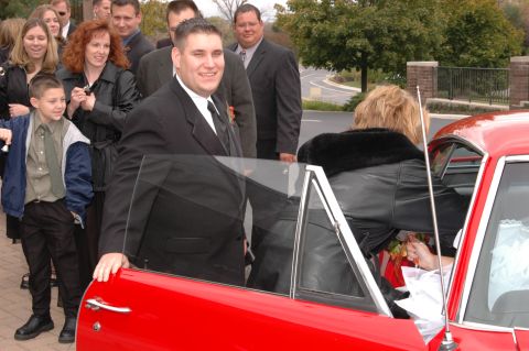 1985 Ford Crown Victoria LTD - A group of people in suits and dresses are gathered around a red car, possibly for a wedding event.
