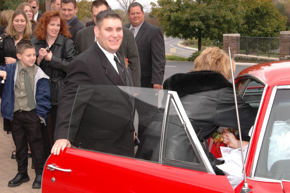 People in formal wear stand next to a red car, with a woman entering the driver's side