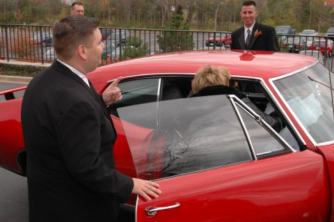 1985 Ford Crown Victoria LTD - A man in a suit opens the passenger door of a vintage red car for a woman. Another man in a suit stands behind them.