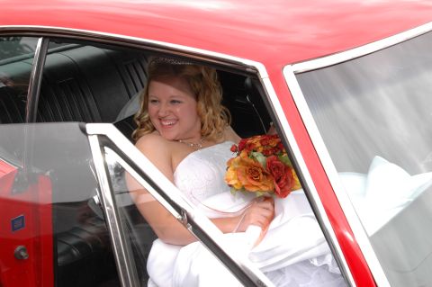 1985 Ford Crown Victoria LTD - Bride smiles while seated in a vintage red car, holding a bouquet of orange and red flowers.