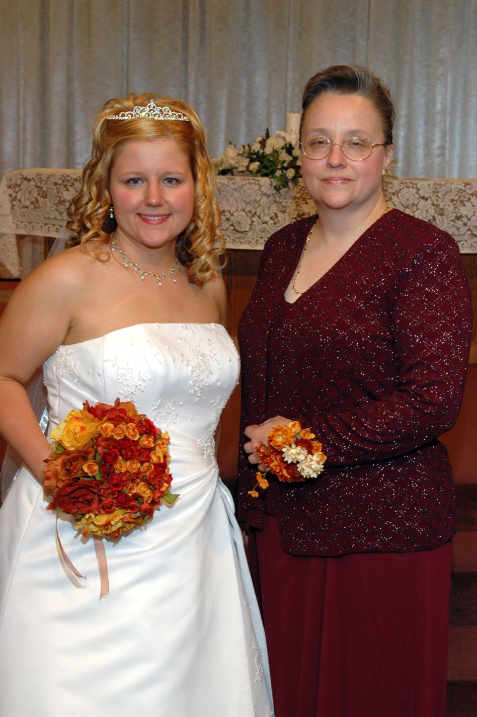 Bride in white dress with tiara and bouquet, and woman in burgundy dress with corsage, pose for a photo
