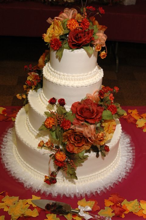 1985 Ford Crown Victoria LTD - A four-tiered white wedding cake is adorned with autumn-colored flowers and ribbons, with a cake server in the foreground.
