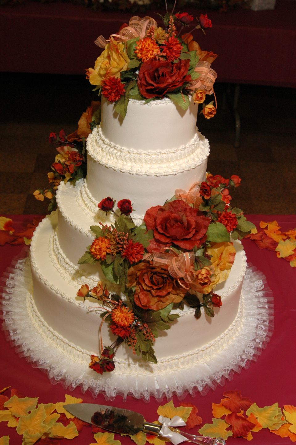 White tiered wedding cake decorated with red, orange, and yellow flowers and ribbons. A cake server lies in front