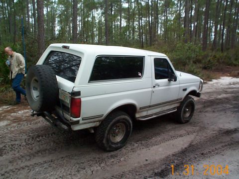 1992 Ford Bronco - A white Ford Bronco is parked on a dirt road surrounded by trees, with a man standing to the left.