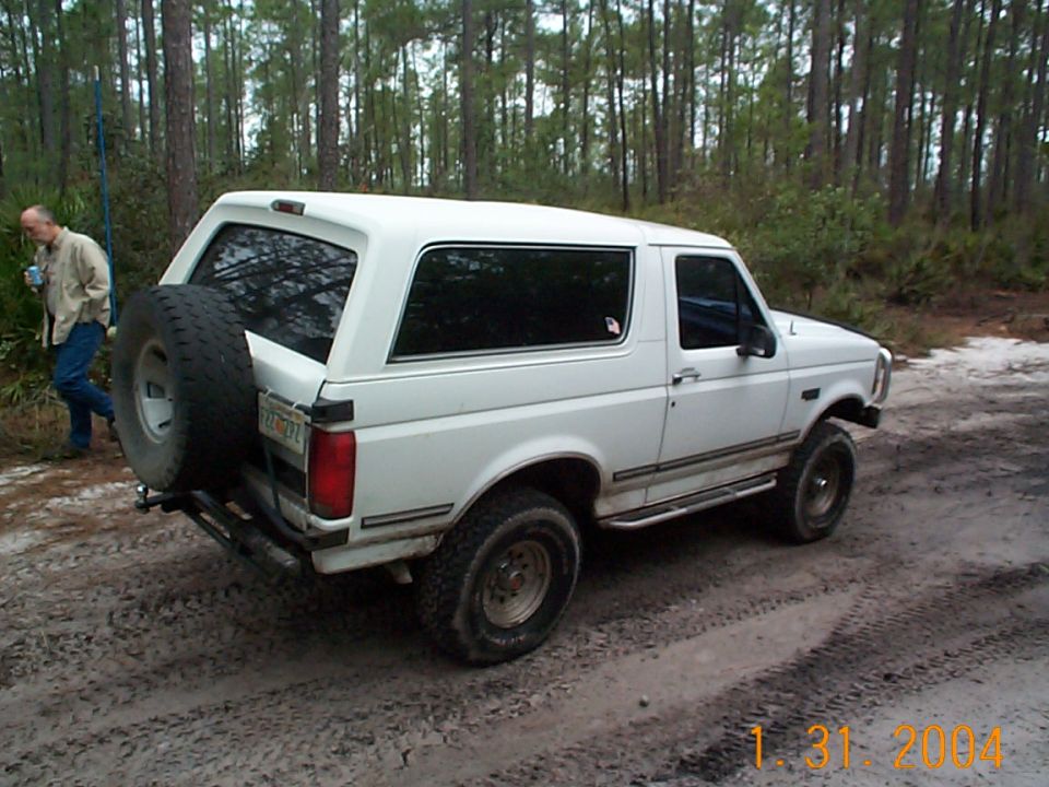 White Ford Bronco parked on dirt road in woods, man standing nearby