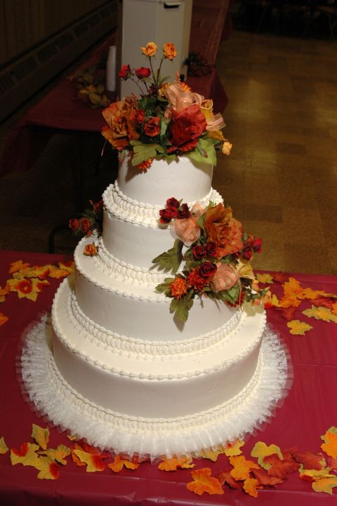 1985 Ford Crown Victoria LTD - A four-tier wedding cake is adorned with floral arrangements in shades of red, orange, and yellow, set against a backdrop of fall leaves.