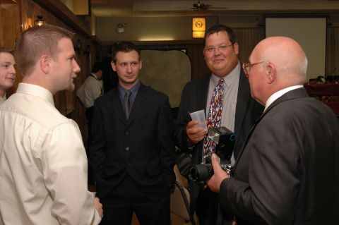 1985 Ford Crown Victoria LTD - Several men in formal attire converse at an indoor gathering, with one man holding a camera and flash unit.