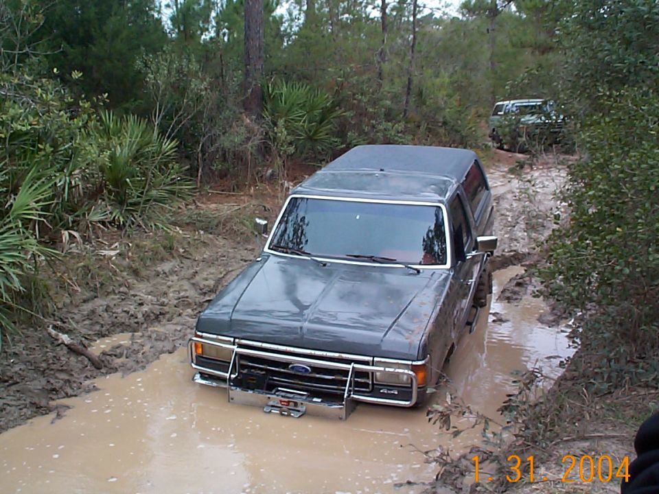 Dark gray Ford Bronco stuck in mud, front bull bar visible, another vehicle in background