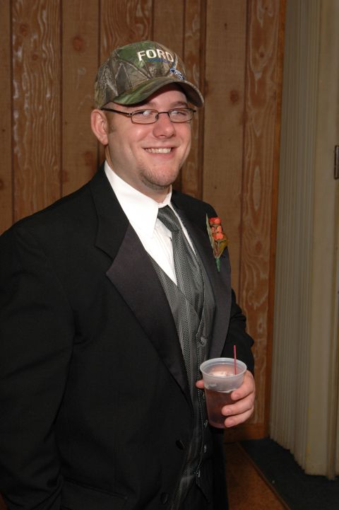 1985 Ford Crown Victoria LTD - Man in tuxedo and camouflage Ford hat holding a drink at an event.