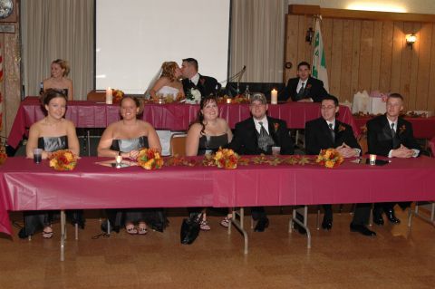 1985 Ford Crown Victoria LTD - A wedding party is seated at a long table during a reception, with a bride and groom kissing in the background.