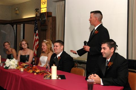 1985 Ford Crown Victoria LTD - A man in a suit gives a toast at a wedding reception, with the bride and groom seated at the head table.