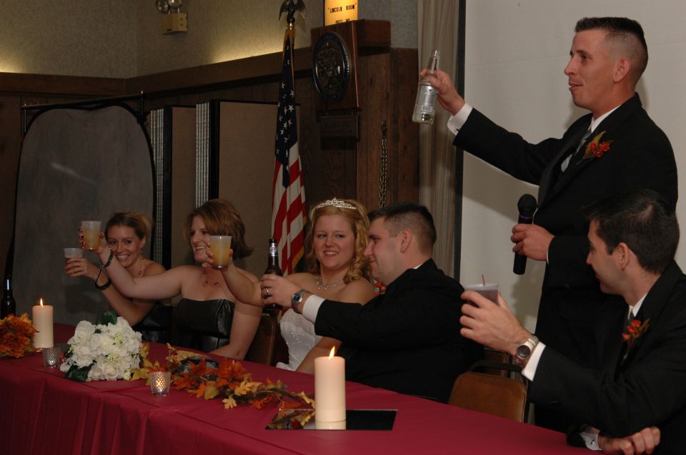 People at a wedding reception table raising drinks. A man in a suit holds a microphone