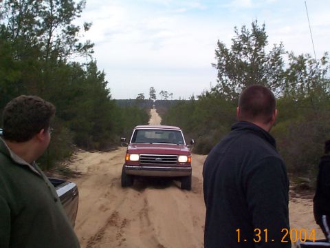 1992 Ford Bronco - A red 1992 Ford Bronco drives down a dirt road, observed by onlookers.