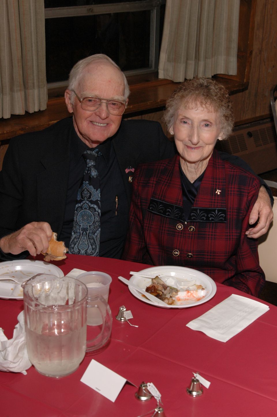 Elderly couple seated at a red table with plates of food and drinks